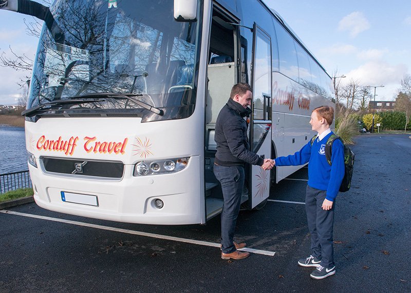 A driver from Corduff Travel shakes hands with a young student in a blue school uniform outside a parked coach, ready for a school trip. The scene is set in a parking area with trees and a river in the background, showcasing a friendly and professional travel experience.