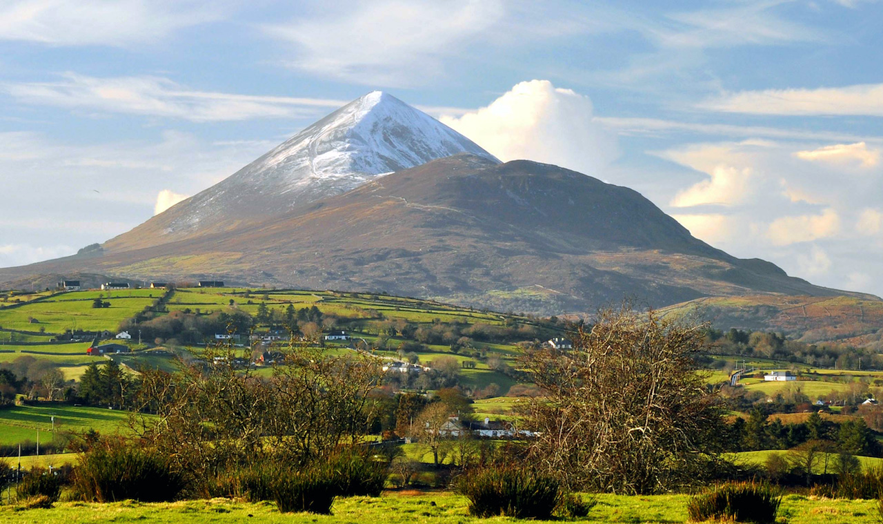 alt="Scenic view of a snow-capped mountain surrounded by lush green fields and scattered houses, under a bright blue sky with wispy clouds."