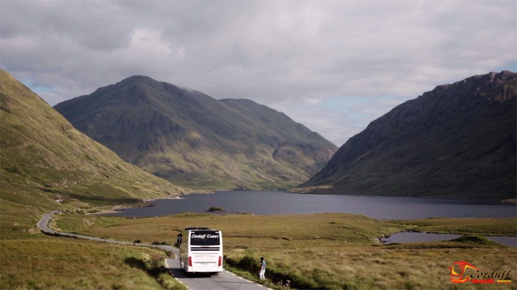Scenic view of a coach traveling along a winding road through lush green mountains, with a serene lake in the background, showcasing the natural beauty of the countryside.