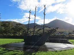 Sculptural representation of a ship with three masts set against a backdrop of mountains and a clear blue sky, located in a park.