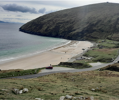 Scenic view of a sandy beach bordered by a lush green hillside, with a winding road leading to the shoreline, under a cloudy sky. The tranquil water reflects the landscape, while a few vehicles are parked near the beach area.