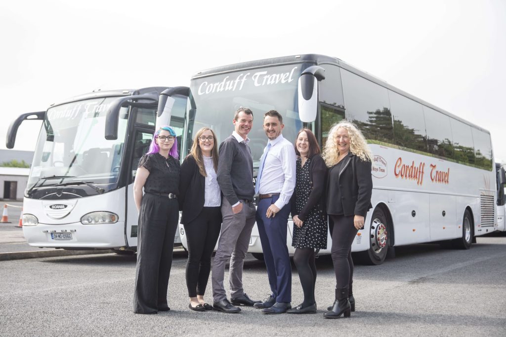 Group of six Corduff Travel team members posing in front of two charter buses, showcasing professional attire and a friendly atmosphere, ideal for promoting group travel services in Ireland.