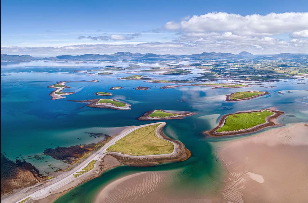Aerial view of a coastal landscape featuring multiple small islands surrounded by turquoise waters, with sandy beaches and lush green vegetation. The scene captures a serene atmosphere with distant mountains and a clear sky, highlighting the natural beauty of the area.