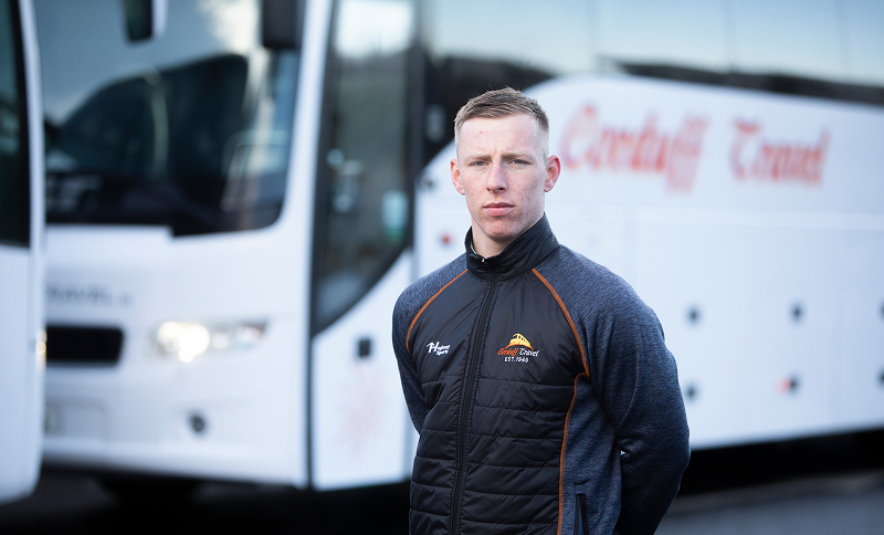 "A young male bus driver stands confidently in front of a coach with the logo of Carduff Travel, wearing a branded jacket."