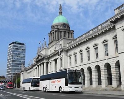 Several white buses parked along the quays beside the Custom House, a grand neoclassical building with a green dome in Dublin, Ireland, under a clear blue sky.