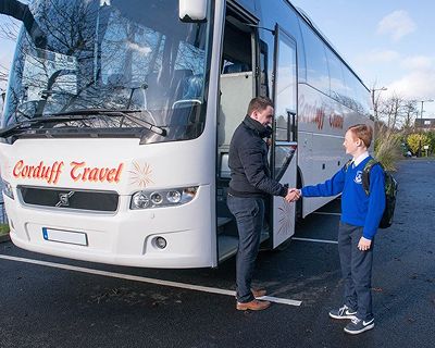 A young student in a blue school uniform shakes hands with a driver in front of a Corduff Travel coach, parked in a lot on a sunny day.