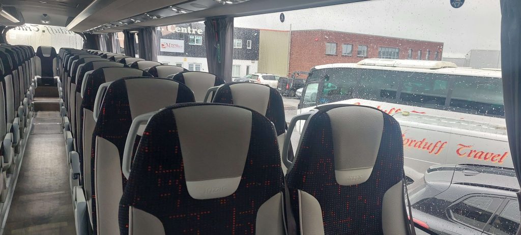 Interior view of a modern travel bus featuring rows of comfortable seats, designed for group transportation. Rain is visible on the windows, with parked vehicles and a building in the background.