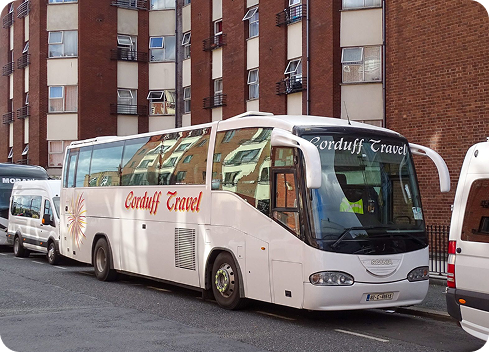 A white coach bus with "Corduff Travel" branding parked on a city street, surrounded by residential buildings and other vehicles.