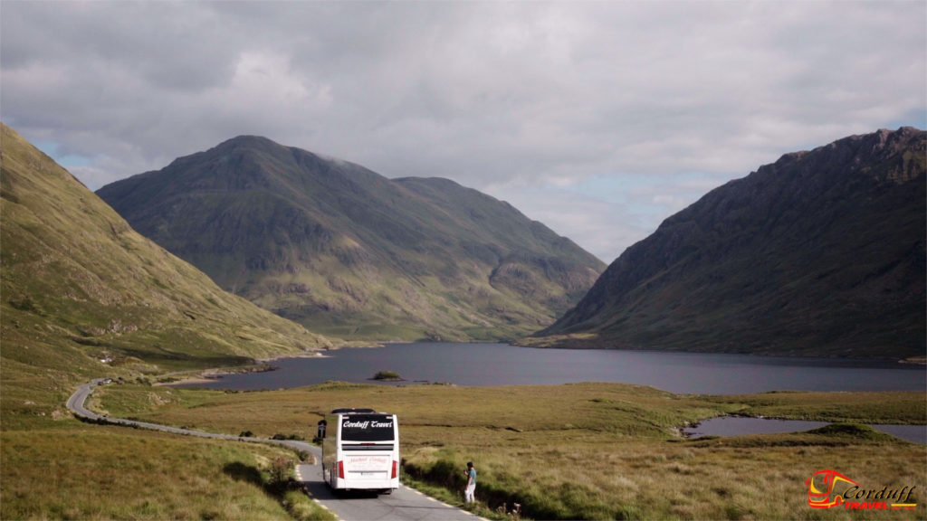 Scenic view of a bus traveling along a winding road in a mountainous landscape, with lush green hills and a tranquil lake in the background, showcasing the beauty of nature in the countryside.