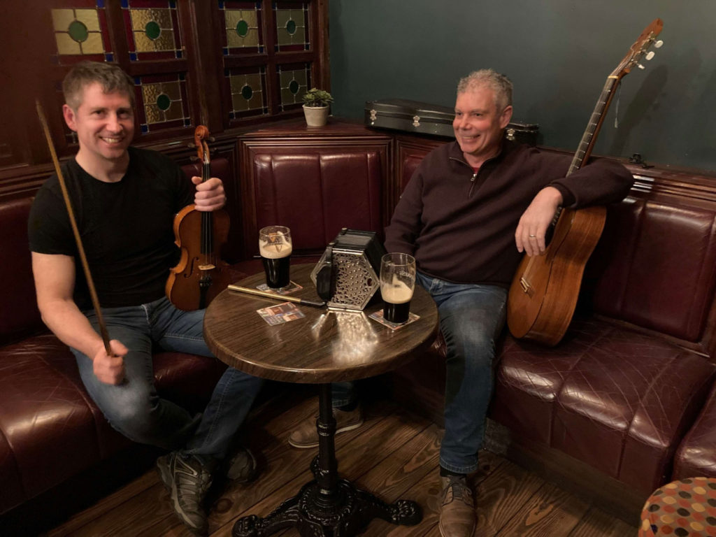 Two musicians enjoying a casual jam session in a cozy pub setting, one holding a violin and bow, while the other leans back with an acoustic guitar. Both have drinks on the table, creating a lively atmosphere for music and conversation.