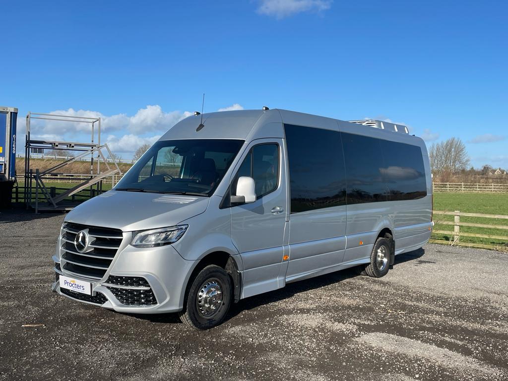 Alt text: Silver Mercedes-Benz Sprinter van parked on gravel with a clear blue sky and green fields in the background.