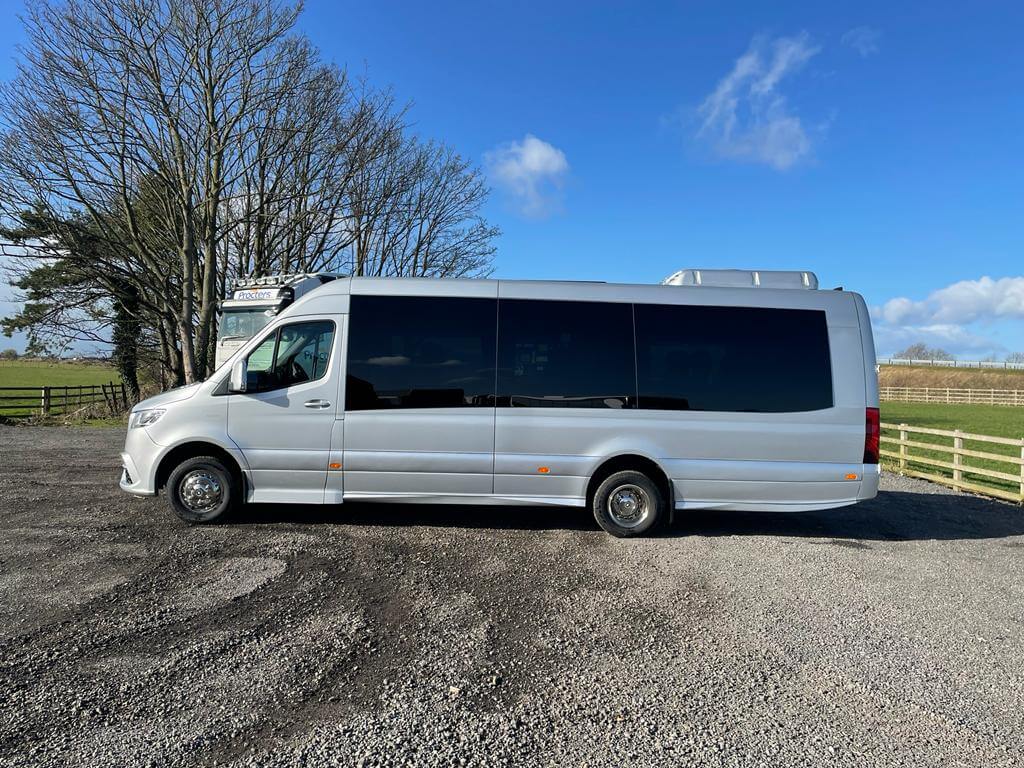A side view of a modern silver passenger van parked on gravel, surrounded by trees and a clear blue sky, showcasing its sleek design and large tinted windows.