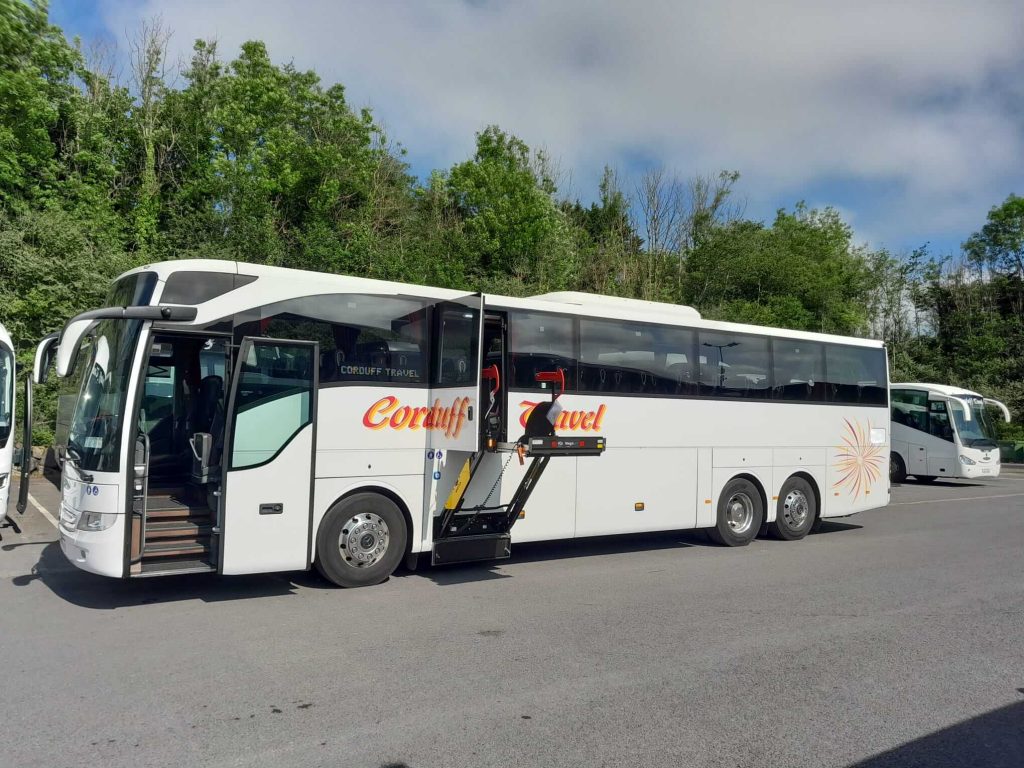 Alt text: Modern coach bus from Corduff Travel parked in a lot, featuring a wheelchair lift for accessibility, surrounded by greenery and other buses.