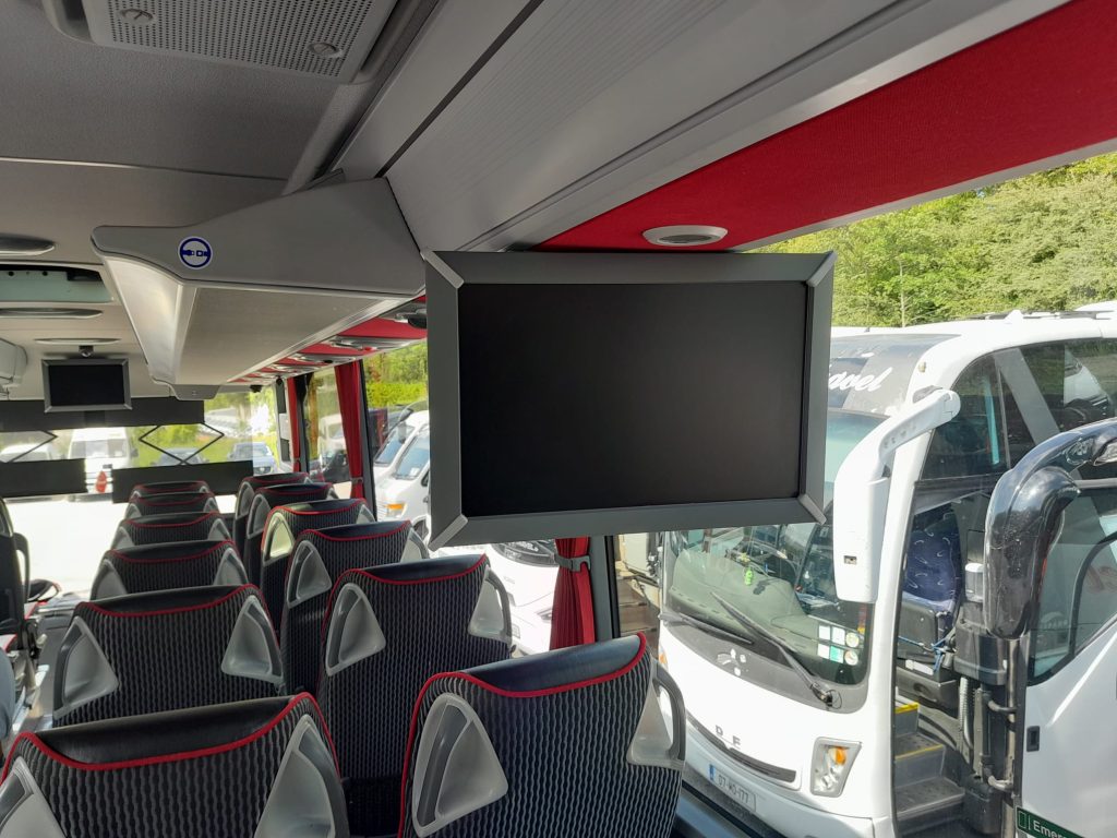 Interior view of a modern bus featuring comfortable seating, overhead storage, and a flat-screen monitor mounted on the ceiling, with a glimpse of other buses parked outside.