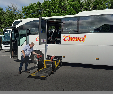 A bus with the word "Travel" on its side parked at a station, featuring a ramp being lowered by a man to assist passengers with mobility issues.