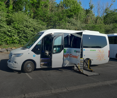 White shuttle bus with open doors parked in a lot surrounded by greenery, promoting travel services.