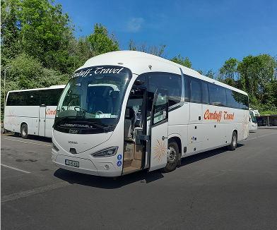 A modern coach bus from Cardiff Travel parked in a lot, with its door open, showcasing its spacious interior and sleek design surrounded by greenery.
