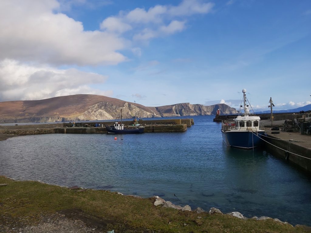 Scenic view of a harbor with fishing boats docked, surrounded by rocky hills and a clear blue sky, highlighting the coastal landscape.