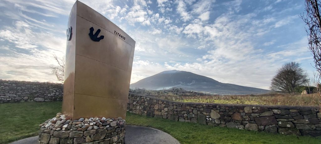 Monument dedicated to the Titanic, featuring a prominent sculpture of a ship's anchor, surrounded by a stone wall and lush green grass, with a scenic view of a mountain and cloudy sky in the background.