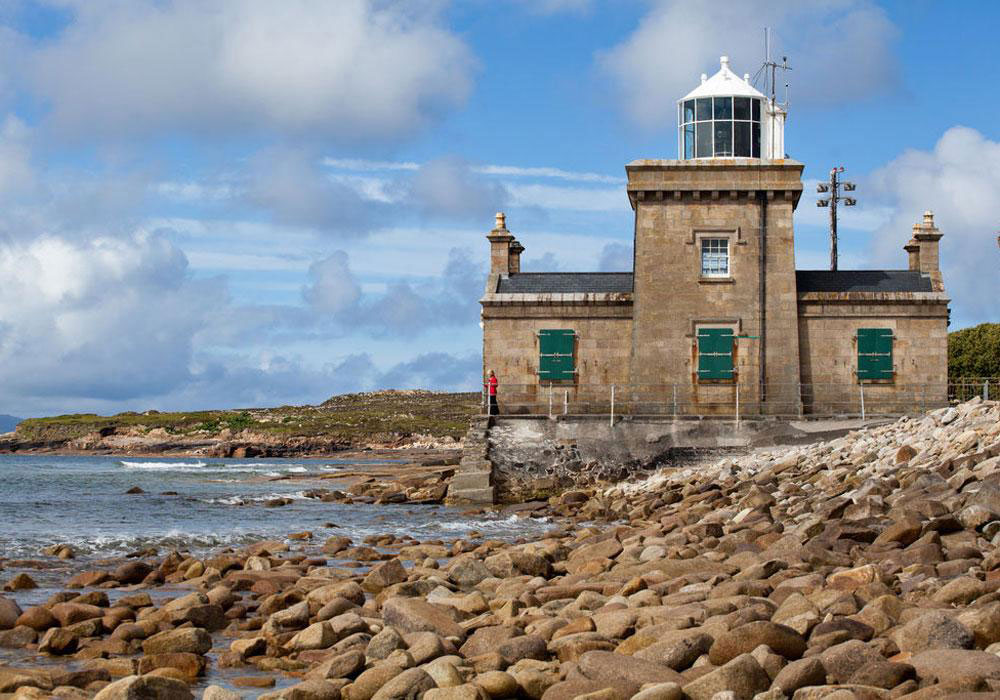 Historic lighthouse on a rocky shoreline with a person in red standing on the steps, surrounded by water and clouds in the background. Ideal for coastal scenery and maritime architecture enthusiasts.