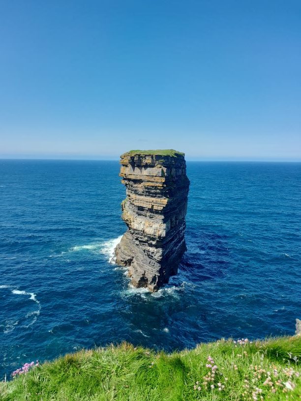 Lone sea stack rising from the ocean with a grassy top, surrounded by blue waters and clear skies, showcasing the natural beauty of coastal landscapes.
