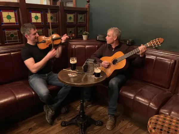 Two musicians perform in a cozy pub setting, one playing the violin and the other strumming a guitar, with two pints of dark beer on the table. The warm ambiance features leather seating and stained glass windows, creating an inviting atmosphere for live music.