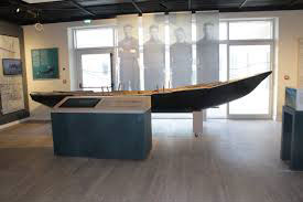 Historic boat displayed in a museum, featuring an interactive exhibit with informational panels and a backdrop of historical figures, highlighting maritime heritage and cultural significance.