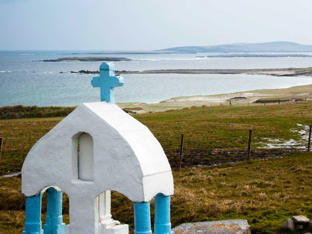 White chapel with a blue cross overlooking the sea and grassy landscape, featuring distant islands under a cloudy sky.