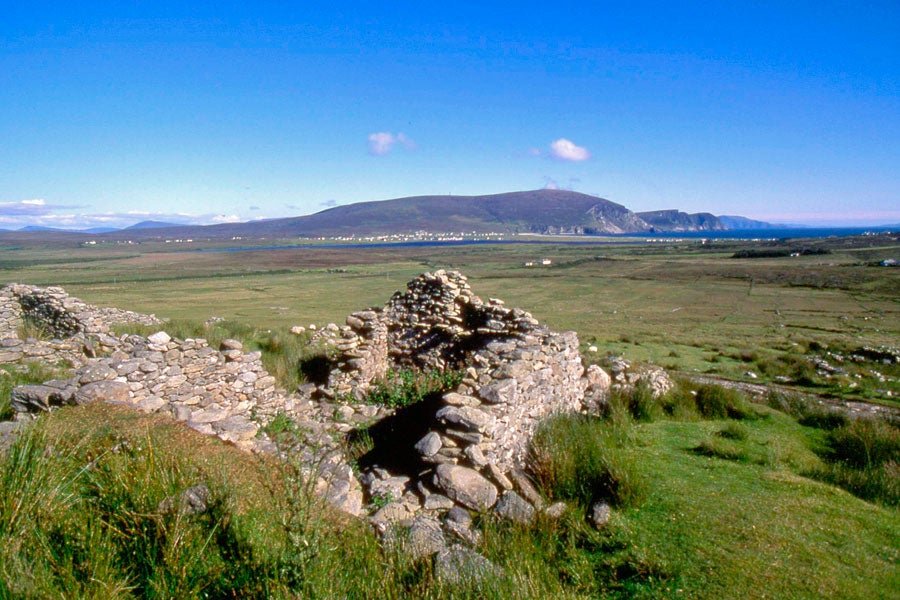 Ruins of an ancient stone structure surrounded by lush green grass, with a panoramic view of rolling hills and a distant coastline under a clear blue sky.