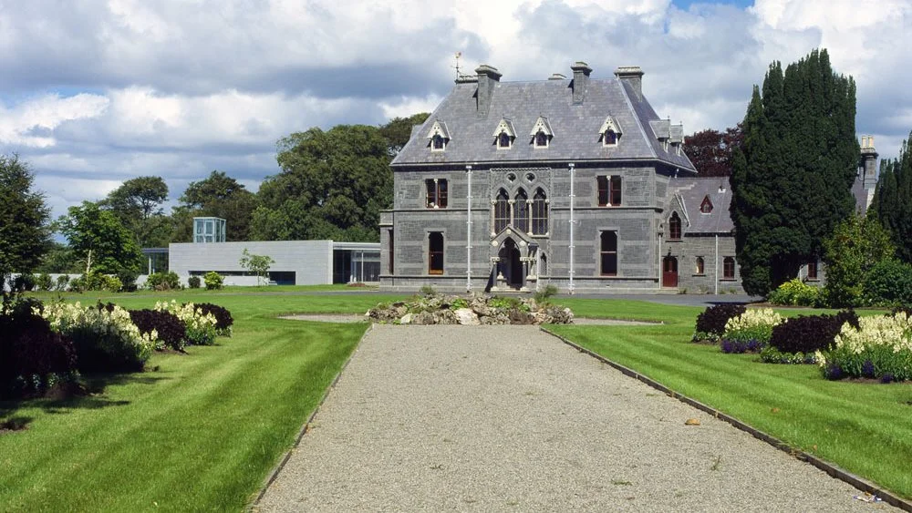 Historic mansion surrounded by lush gardens and a gravel pathway, showcasing a blend of traditional and modern architecture under a partly cloudy sky.